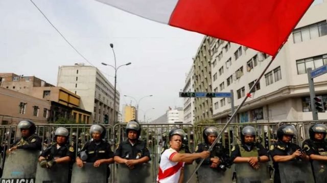 Seorang pria mengibarkan bendera Peru, sementara petugas polisi berjaga selama protes setelah Kongres menyetujui pencopotan Presiden Pedro Castillo, di Lima, 7 Desember 2022 [Alessandro Cinque / Reuters]