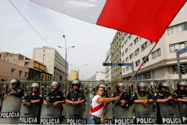 Seorang pria mengibarkan bendera Peru, sementara petugas polisi berjaga selama protes setelah Kongres menyetujui pencopotan Presiden Pedro Castillo, di Lima, 7 Desember 2022 [Alessandro Cinque / Reuters]